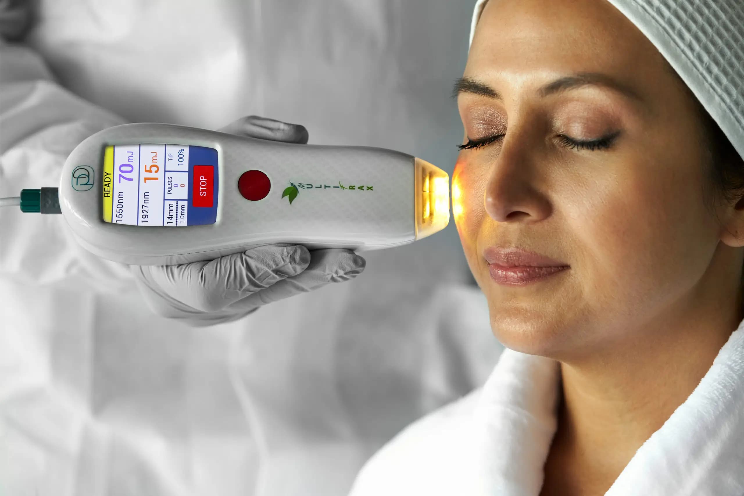 A woman receiving a MultiFrax laser facial, the handpiece resting at her cheek with its 1550nm and 1927nm display active